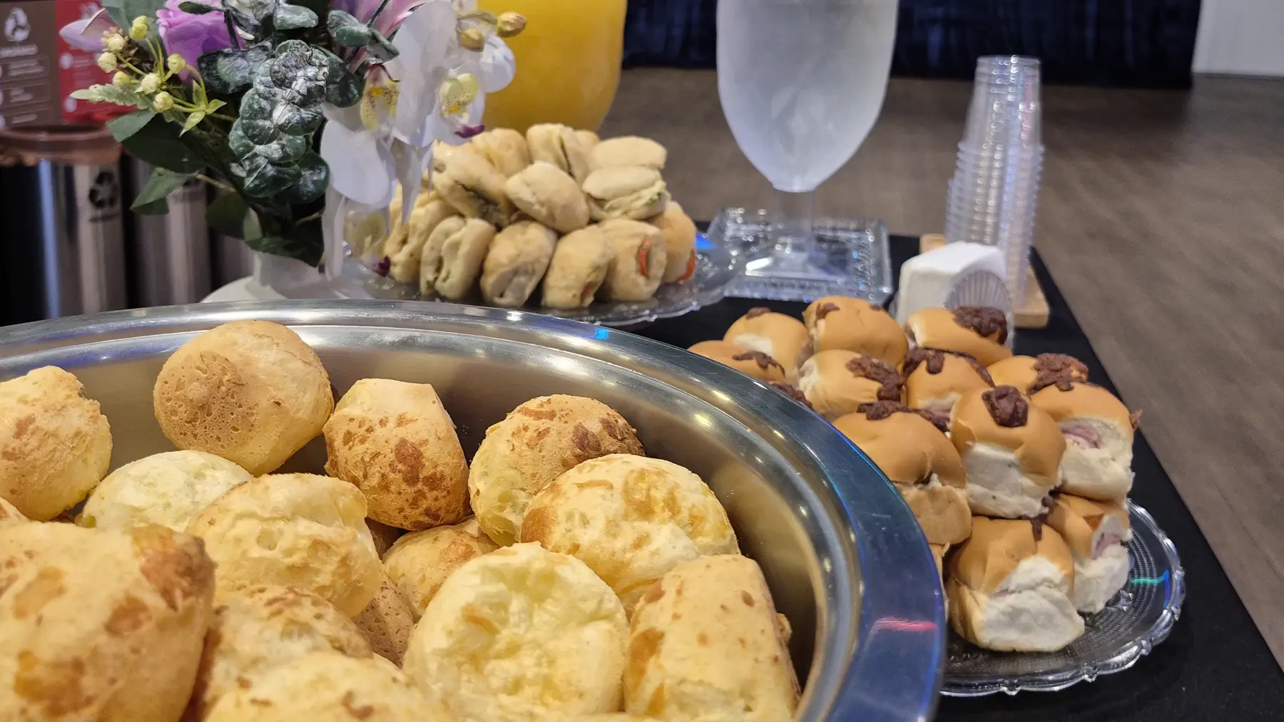 Travessa de pão de queijo, mini-sanduíches e arranjo de flores numa mesa de coffee break montada